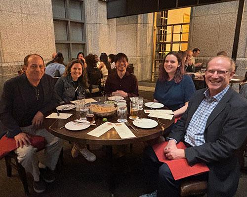 Five people seated around a round restaurant table pose for a photo with other diners visible in the background
