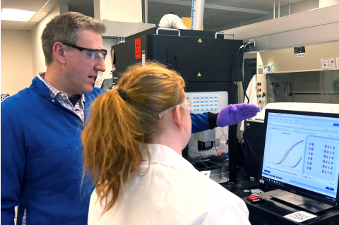 Professor Charlie Mace and a student collaborate in a laboratory, surrounded by scientific equipment and research materials